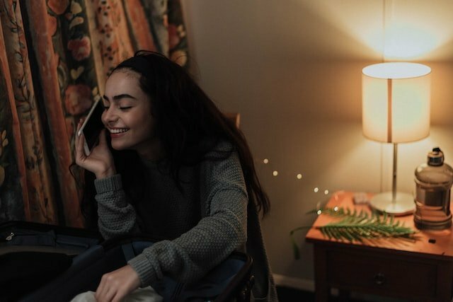 A picture of a young lady speaking on her mobile phone beside a lamp that is luminating on a table beside her with a leaf and a water bottle.