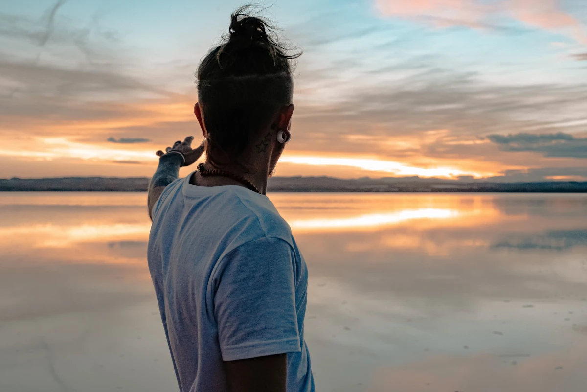 A man with tattoos and a man bun on a beach with his arm extended. The picture is from the back as he faces out to the sand and water.