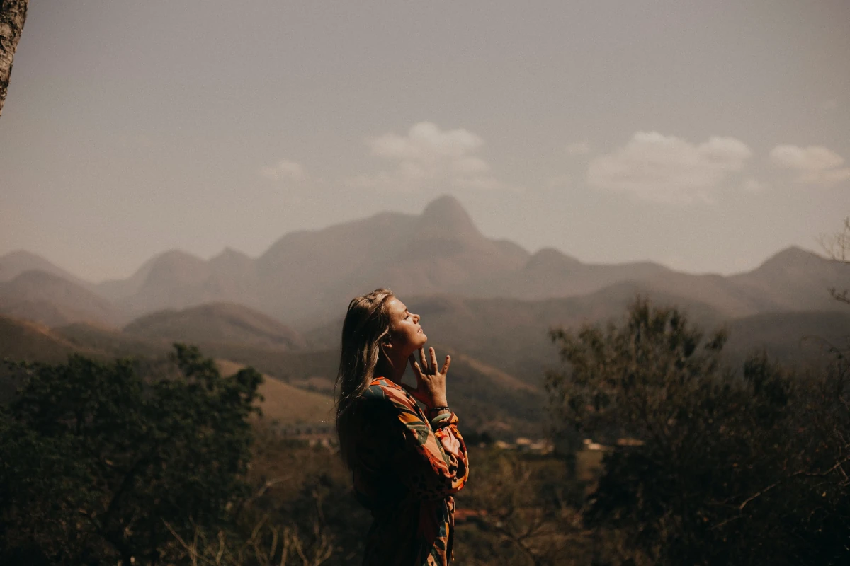 A lady facing to the right with her hands together with fingers spread under her chin. Her eyes are closed and she has long hair. Behind her is a valley and a mountain.